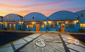 Blue buildings with arched roofs and lit windows under a sunset sky, with a mural on the pavement in front.