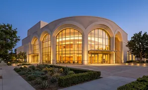 Modern beige building with large arched windows and warm interior lighting at dusk.