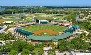 Aerial view of a baseball stadium surrounded by trees and residential areas under a clear blue sky.