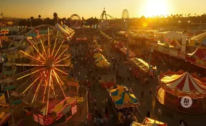 Aerial view of a bustling carnival at sunset, with illuminated Ferris wheels, rides, and crowds of people.