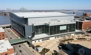 Aerial view of a large modern arena with glass facades, a curved roof, and a pedestrian bridge connecting it to a nearby building.
