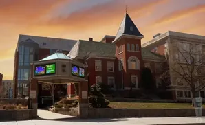 An old brick building with a tower and a modern glass addition is under a colorful sunset sky.