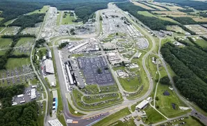 Aerial view of a race track with grandstands, parking lots full of cars, and surrounding trees.