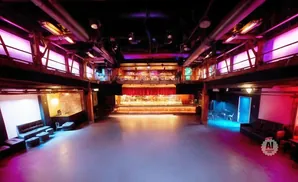 Empty concert venue with stage, bar, and seating under purple and pink lighting.