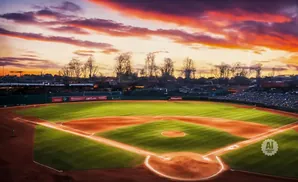 A baseball field at sunset with a dramatic orange and purple sky.
