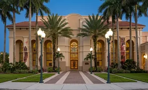 Exterior of a tan building with arched windows and red tile roof, flanked by palm trees and lit by ornate lampposts.