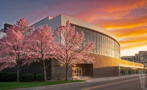A modern building with a curved glass facade and brick base, framed by blooming pink cherry trees at sunset.