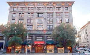 An ornate, multi-story stone building with many windows, framed by trees and a street scene with cars.