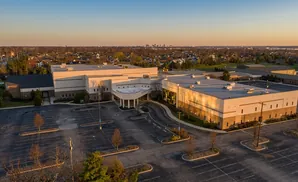 Aerial view of a modern church complex at sunset, with a large parking lot in the foreground and a distant cityscape.