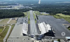Aerial view of a drag racing strip with grandstands and a large parking lot surrounded by trees and a distant golf course.