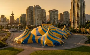 A circus tent with yellow and blue stripes sits in front of a city skyline at sunset.