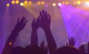Silhouetted hands are raised in the air at a concert with purple and yellow stage lights.