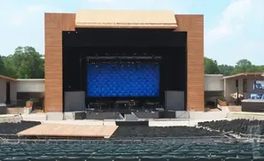 An outdoor amphitheater stage with a large blue LED screen, flanked by speakers and wooden pillars, facing rows of empty seats.