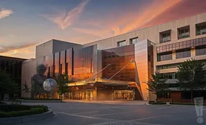 A modern building with glass facade reflects a colorful sunset, with a large sculpture of a person holding a sphere in front.