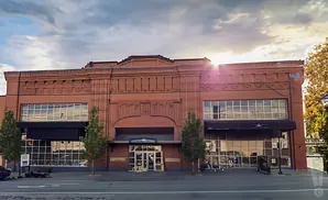 A brick building with large windows, trees, and the sun shining.