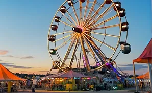 A Ferris wheel illuminated at dusk at a fairground, with tents and rides below.