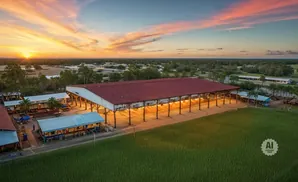 An aerial view of a long, red-roofed barn illuminated by warm lights at sunset.