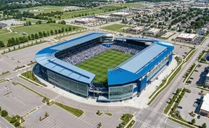 Aerial view of a soccer stadium with blue roofing, surrounded by parking lots and residential and commercial buildings.