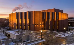 Sunset illuminates a modern brick building with vertical windows, steam rises from a chimney.