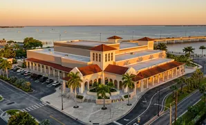 An aerial view of a large, tan building with a red tile roof, resembling a theater or convention center, with palm trees and a parking lot.