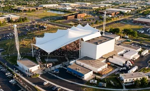 Aerial view of an outdoor amphitheater with a large white tent, crowded seating, and surrounding industrial buildings and parking lots.