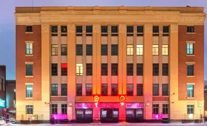 A tan and brick building with lit windows and pink neon signs glows at dusk.