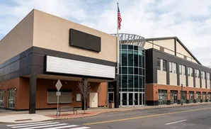 A modern brick and tan building with a curved glass entrance, marquees, and an American flag.