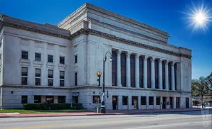A grand stone building with classical columns stands under a bright blue sky with a radiant sun.