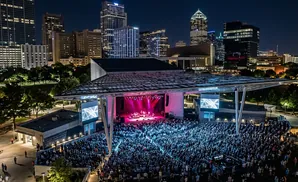 An outdoor concert at night with a band on stage, a large crowd, and a city skyline in the background.