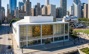 Modern white building with large glass windows displaying artwork, set against a cityscape and park.