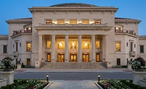 Neoclassical building with columns, lit windows, and ornate details, flanked by lush landscaping.