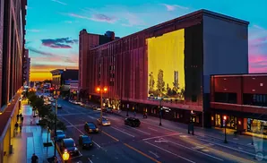 A city street at dusk with a mural of trees and towers on a large brick building.