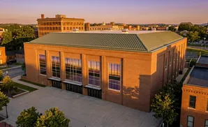 A large brick building with a green roof and tall windows reflecting the sunset, viewed from above.