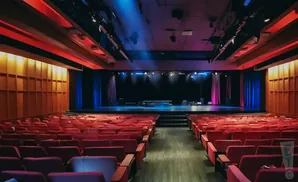 An empty theater with red seats, a stage with blue and pink curtains, and overhead stage lighting.
