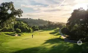 Golfer on a lush green golf course with rolling hills and trees under a hazy, sunlit sky.