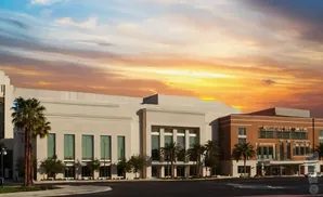 A modern building with a tan facade and large windows, surrounded by palm trees, under a dramatic sunset sky.