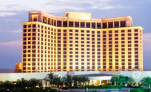 A tall, curved hotel building at dusk, lit by warm interior lights, with palm trees in the foreground.