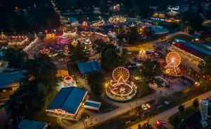 Aerial view of a lively carnival at night, with Ferris wheels, rides, tents, and crowds illuminated by festive lights.