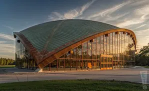 A modern building with a curved, green roof and glass walls, reflecting the sunset.