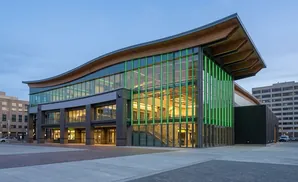 Modern building with a wavy wooden roof, green-lit glass facade, and a paved plaza.
