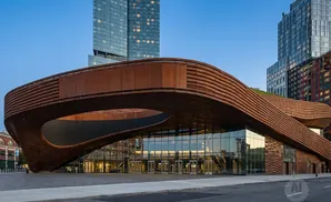 Modern building with curved, copper-colored facade and glass storefronts, next to tall buildings.