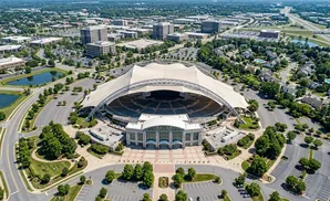 Aerial view of an amphitheater with a large white canopy, surrounded by trees, parking lots, and buildings.