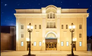 An illuminated theater building at night with a starry sky above and two ornate street lamps in the foreground.