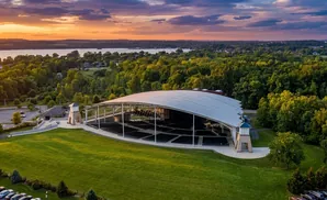 Aerial view of an outdoor amphitheater with a large white roof and rows of seats, surrounded by green trees and a lake at sunset.