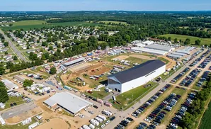 Aerial view of a fairground with buildings, parking lots, RVs, and a Ferris wheel under a blue sky.