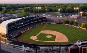 Aerial view of a baseball stadium at dusk with lights on, featuring green outfield and brown infield.