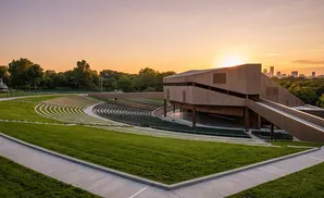 Outdoor amphitheater with tiered seating, a modern stage, and a city skyline at sunset.