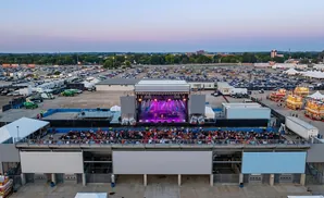 Aerial view of an outdoor concert stage with a band performing, facing a seated audience, with a large parking lot and fairgrounds in the background.
