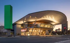 A modern convention center with a large, curved roof and a tall green LED tower in front.