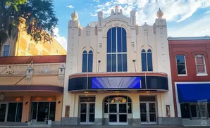 Art deco theater with a large marquee screen and multiple arched windows, under a cloudy blue sky.